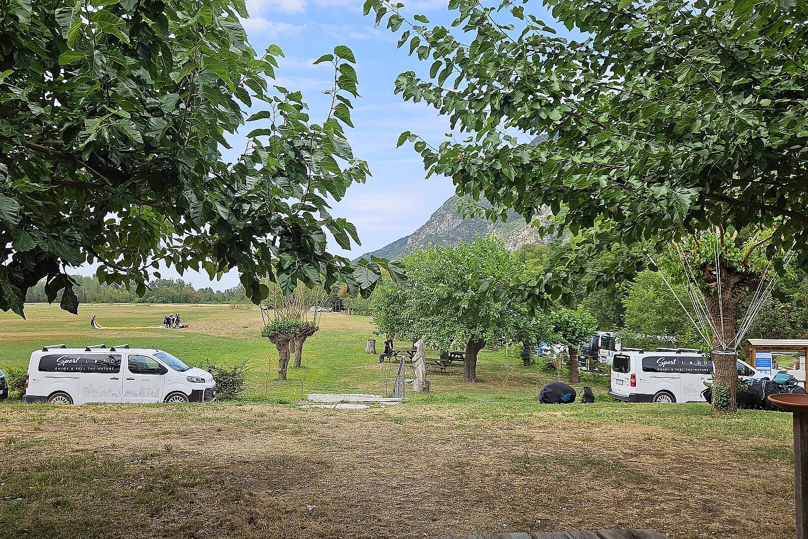Wiese mit mehreren Fahrzeugen, Bäumen und verstreuten Ausrüstungsgegenständen.