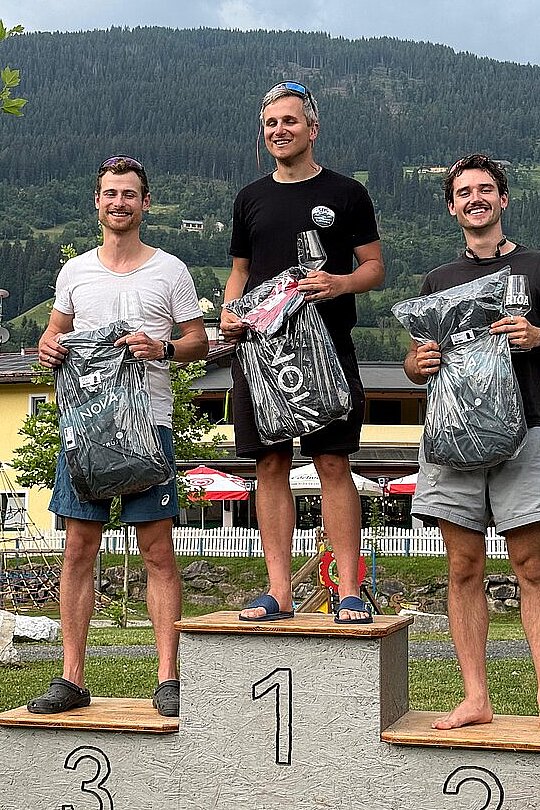 Drei Männer auf dem Siegerpodest mit Preisen vor einer Bergkulisse.
