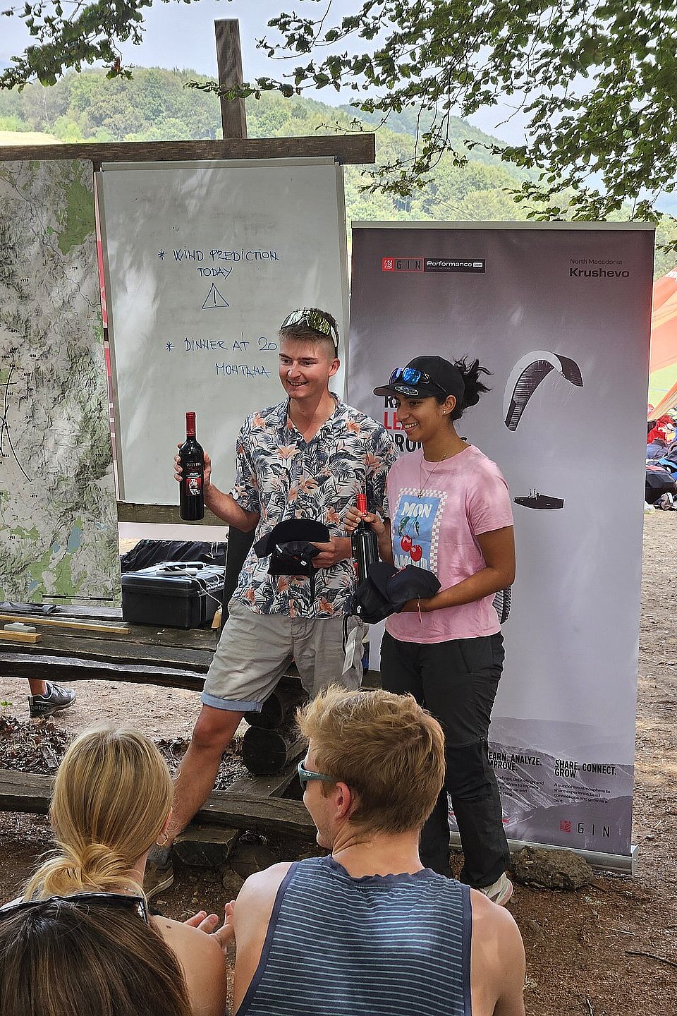 Zwei Personen stehen vor einer Tafel mit Karte und Bannern und halten Flaschen in der Hand.