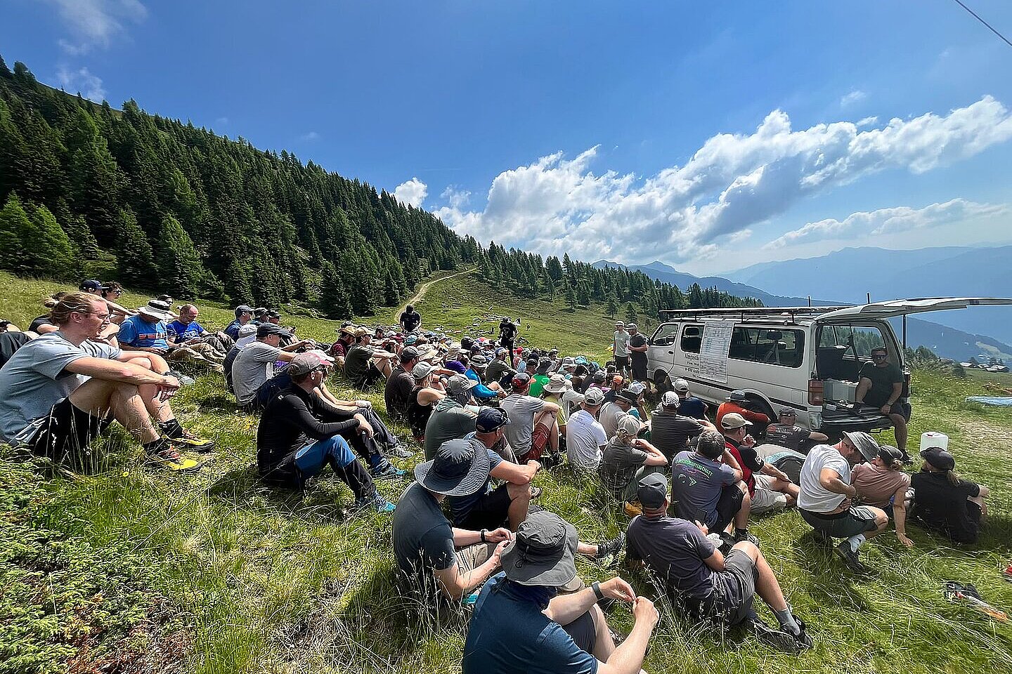 Viele Menschen sitzen am Startplatz auf einer Wiese neben einem Fahrzeug und schauen zu.