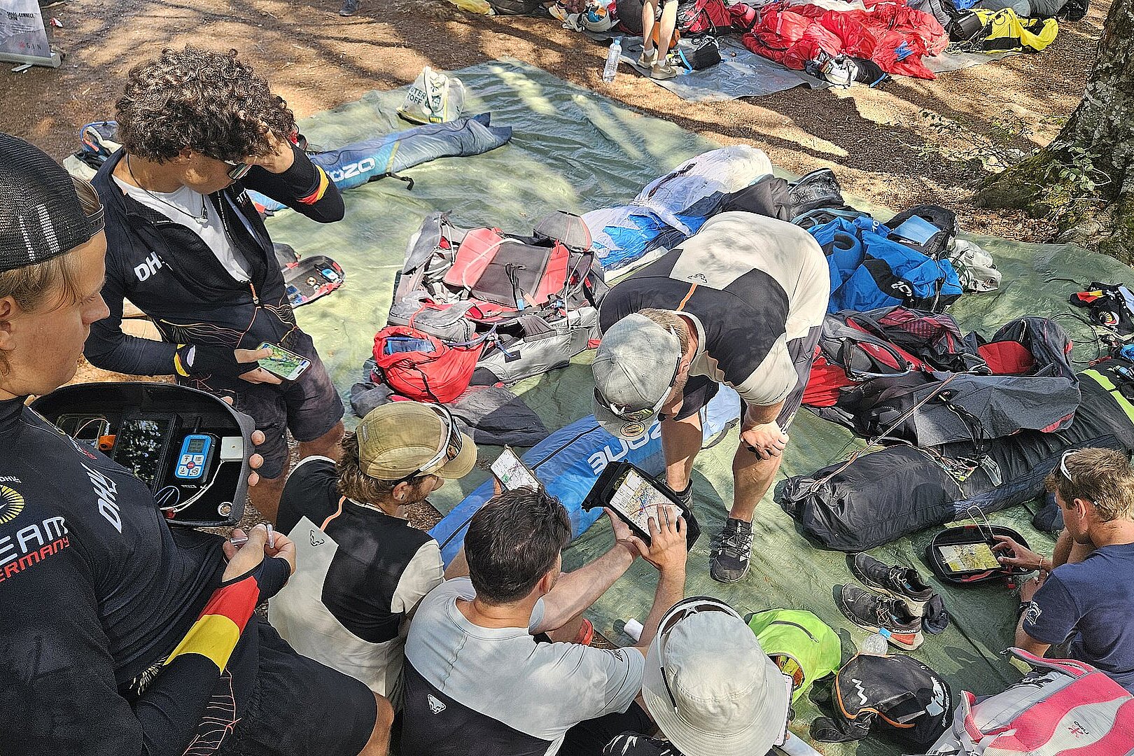 Gruppe von Piloten sitzt im Wald um Karten und Geräte, Ausrüstung liegt ausgebreitet.