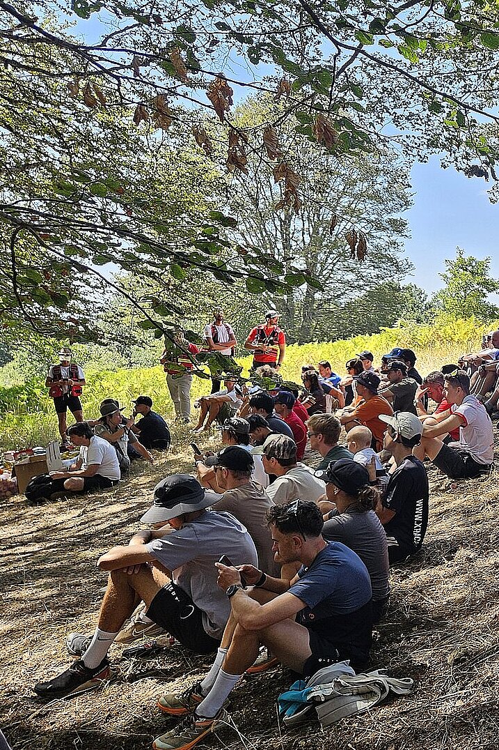 Viele Teilnehmende sitzen im Schatten unter Bäumen und warten auf den Start.