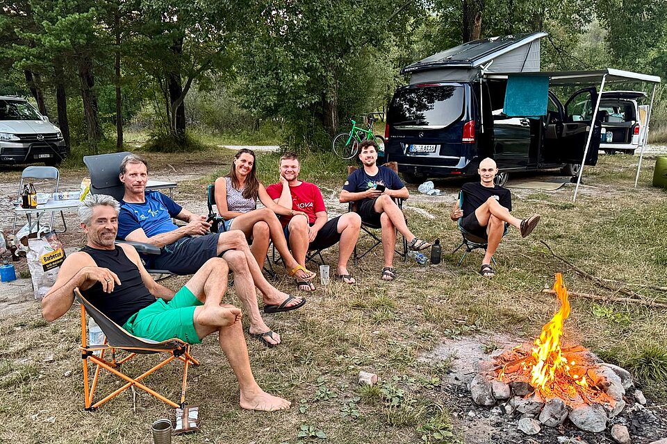 Sechs Personen sitzen im Freien auf Campingstühlen am Lagerfeuer neben Transportern.