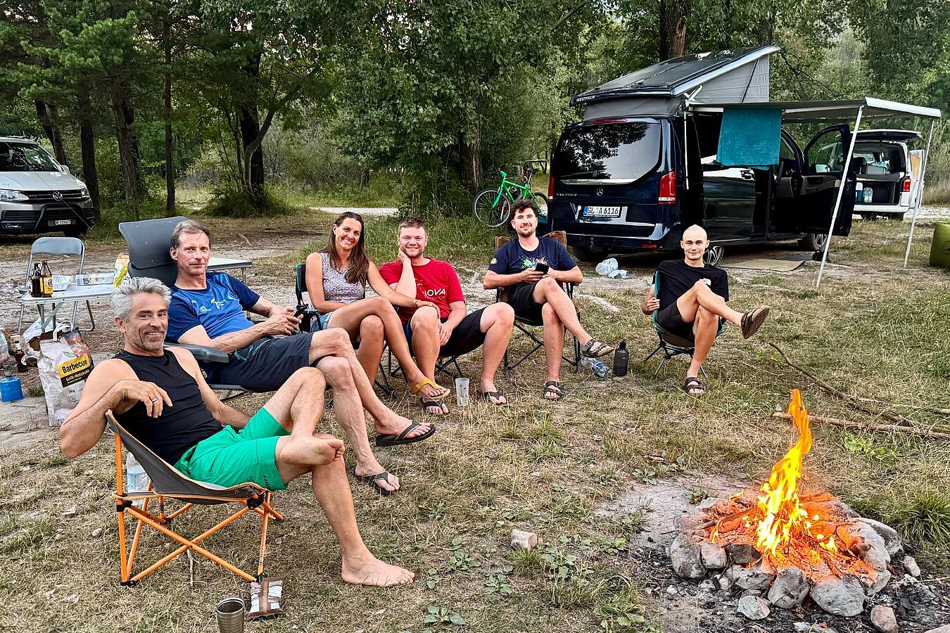Sechs Personen sitzen im Freien auf Campingstühlen am Lagerfeuer neben Transportern.