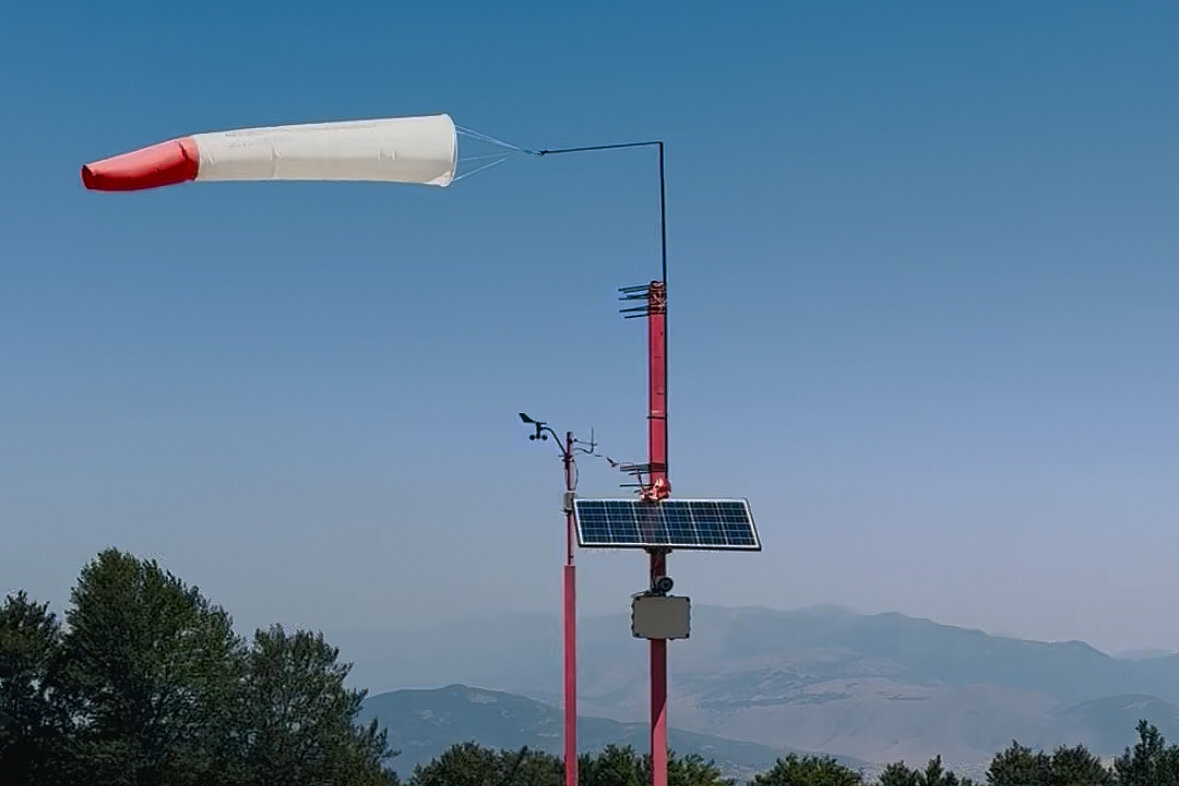 Windfahne an einem Mast vor Bergen und Bäumen