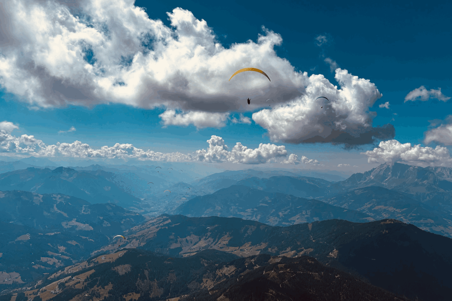 Paraglider unter großen Wolken über einem weiten Bergtal, weitere Schirme in der Ferne.