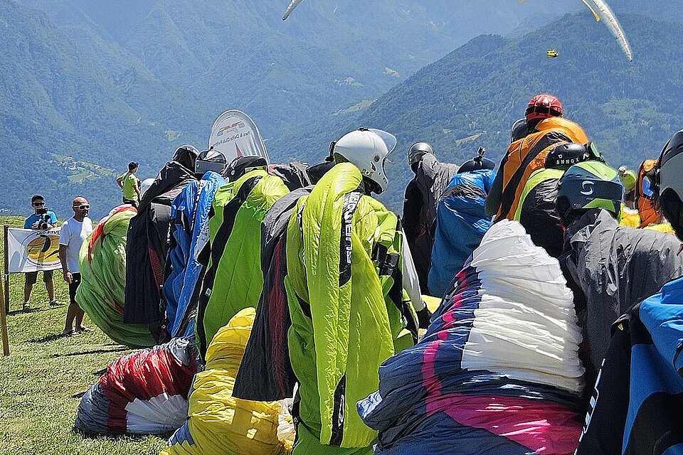 Piloten warten am Startplatz mit ausgebreiteten Gleitschirmen vor Bergkulisse.