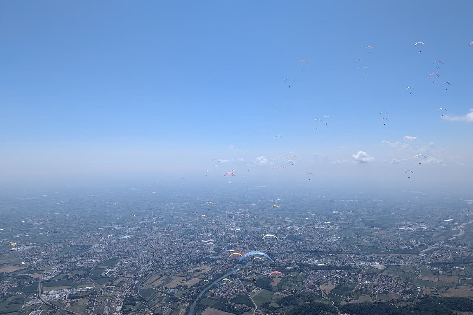 Zahlreiche Gleitschirme schweben über einer weiten Stadtlandschaft im blauen Himmel.