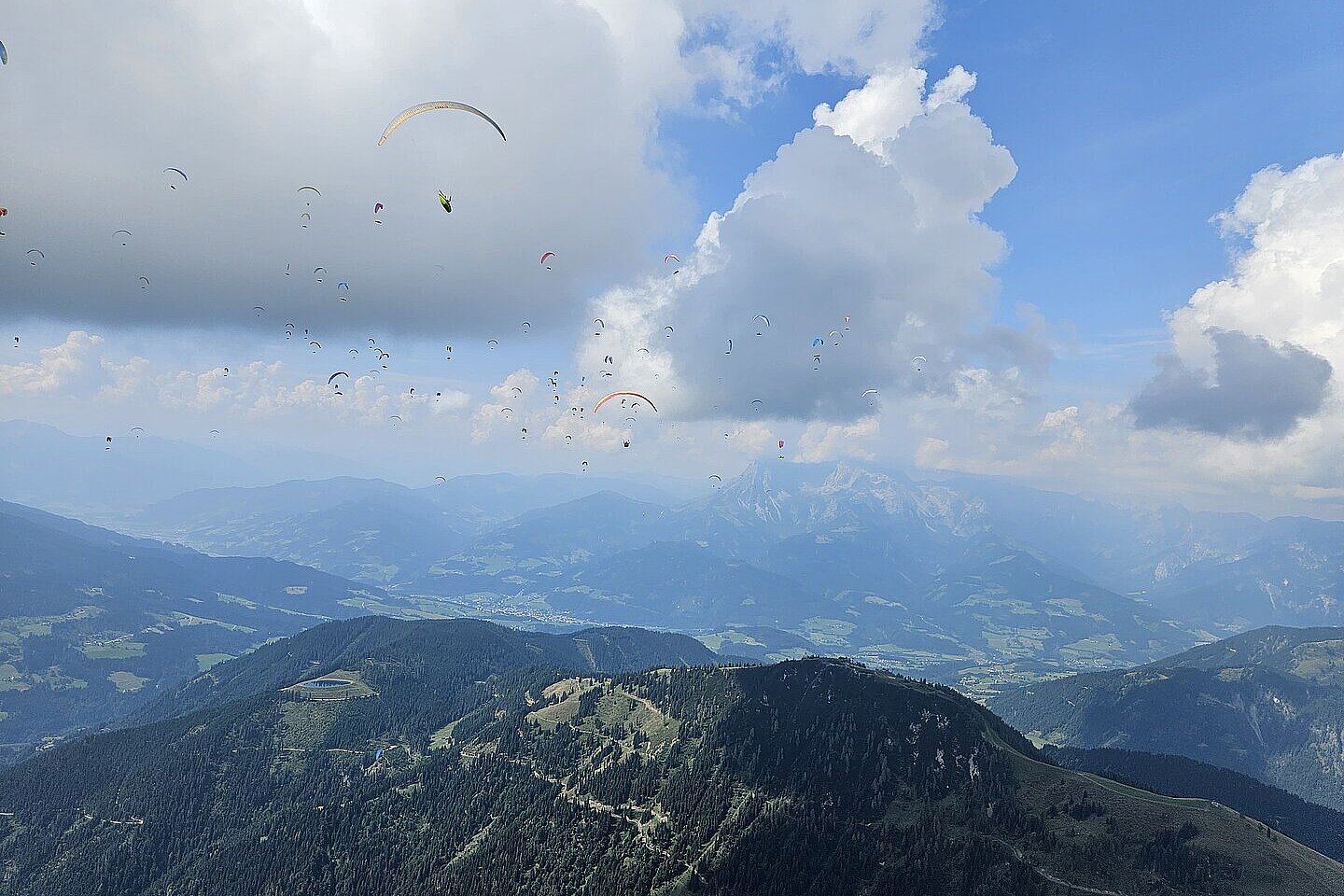 Viele Gleitschirme fliegen über Bergkämme und ein weites Tal mit Wolken und Fernsicht.