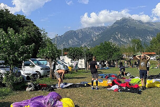 Piloten packen Gleitschirme auf einer Wiese mit Autos, Bäumen und Bergen im Hintergrund.