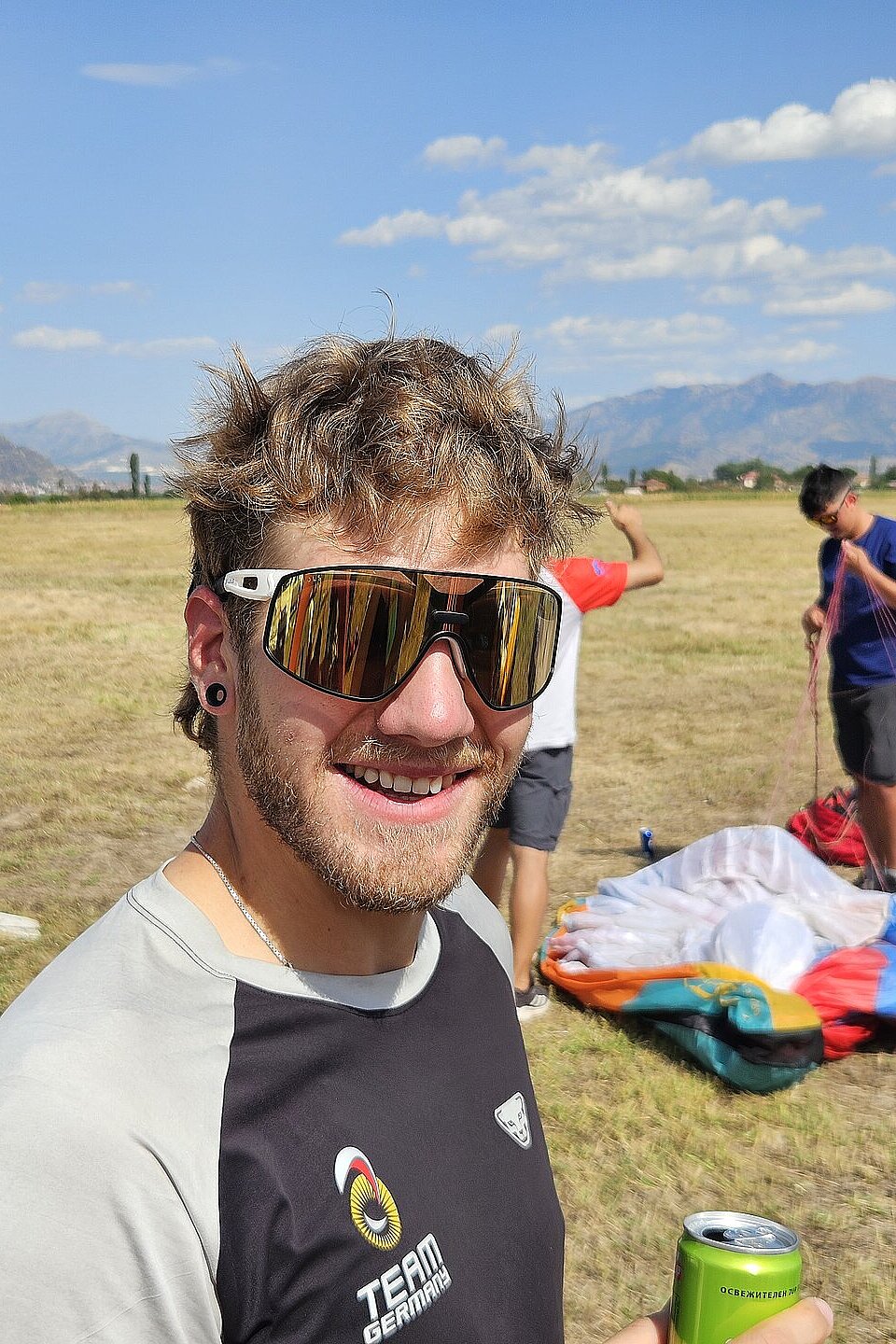 Mann mit Sonnenbrille lächelt in die Kamera, hinter ihm packen zwei Personen einen Gleitschirm zusammen.