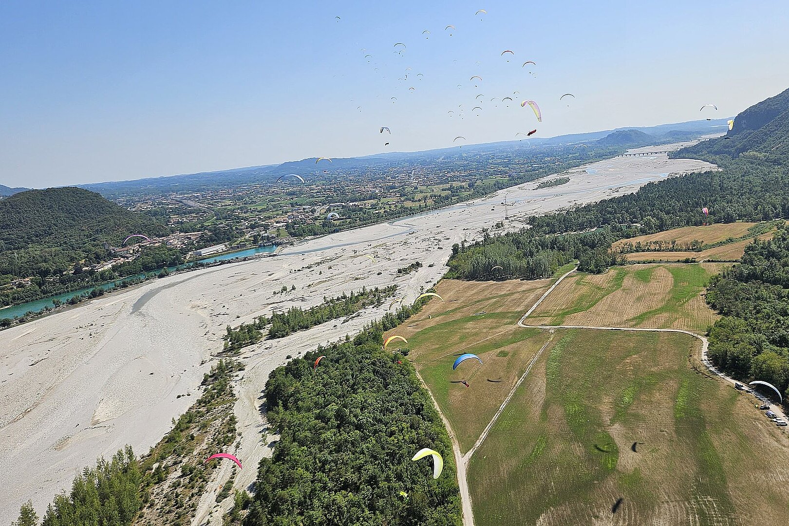 Viele Gleitschirme schweben über einem breiten Flussbett und einer grünen Ebene.