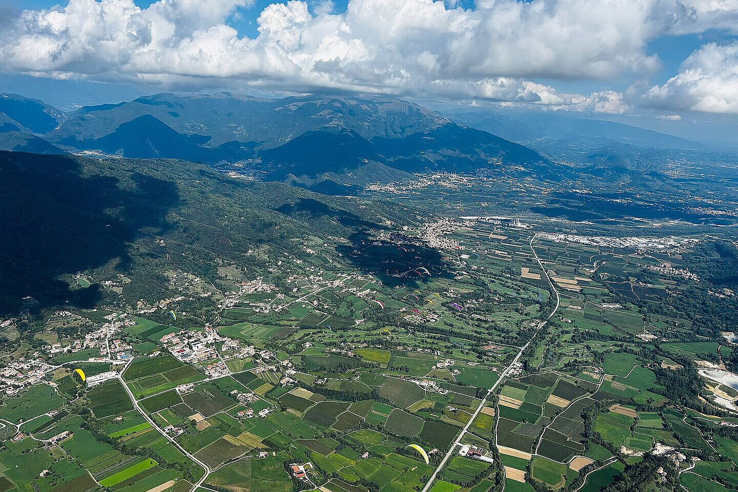 Mehrere Gleitschirme fliegen über einer grünen Tal- und Hügellandschaft mit Wolken am Himmel.