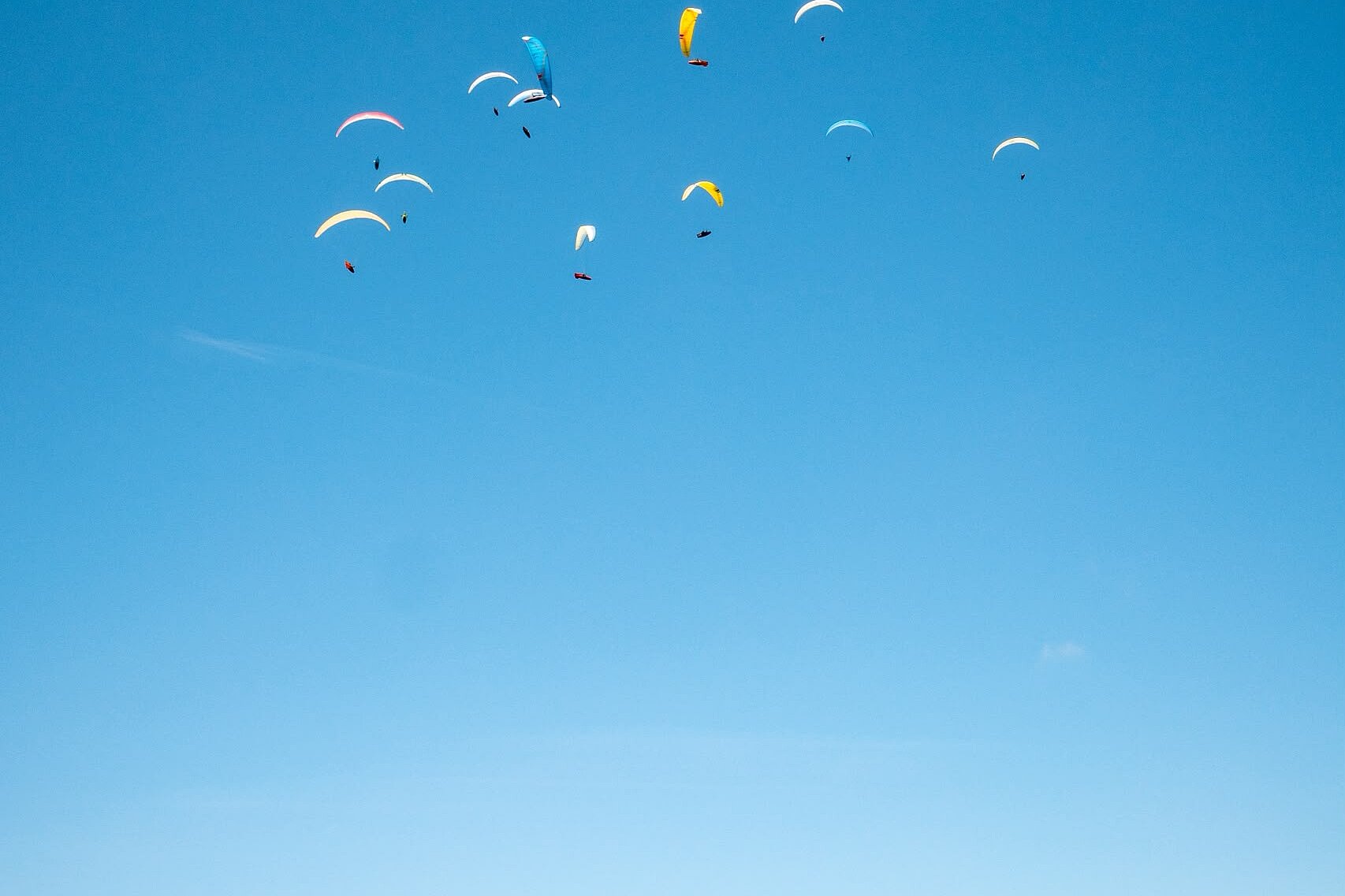 Mehrere Gleitschirme fliegen hoch am blauen Himmel über einer Berglandschaft.