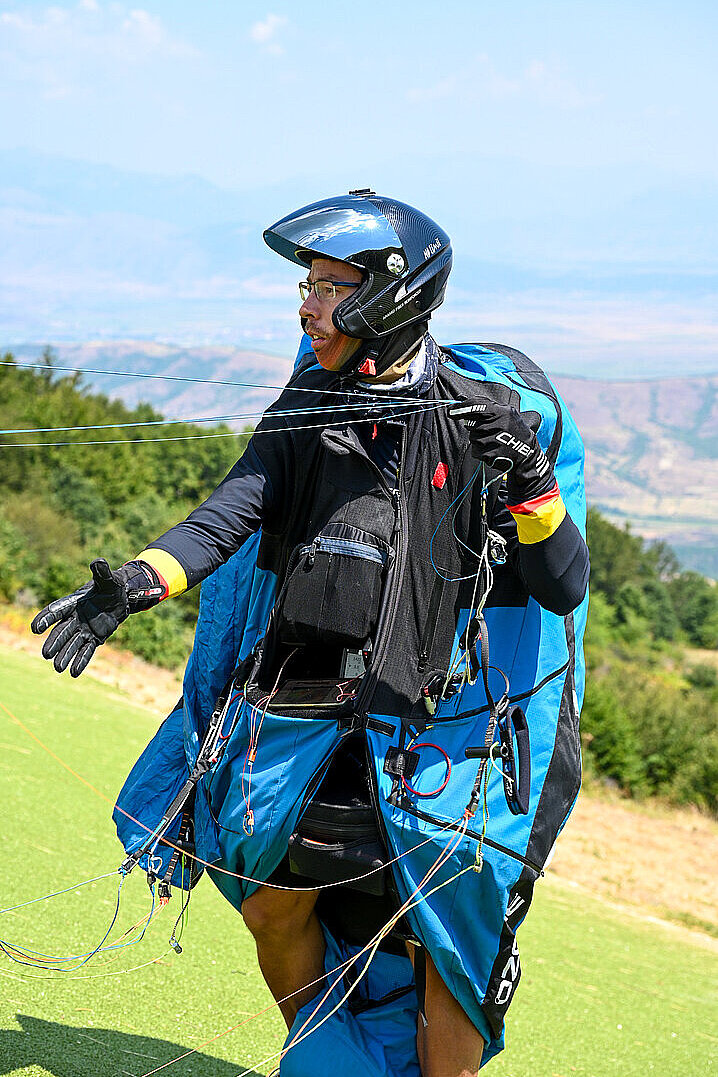 Gleitschirmpilot mit Helm und Gurtzeug steht auf einer Wiese und hält die Leinen in der Hand.