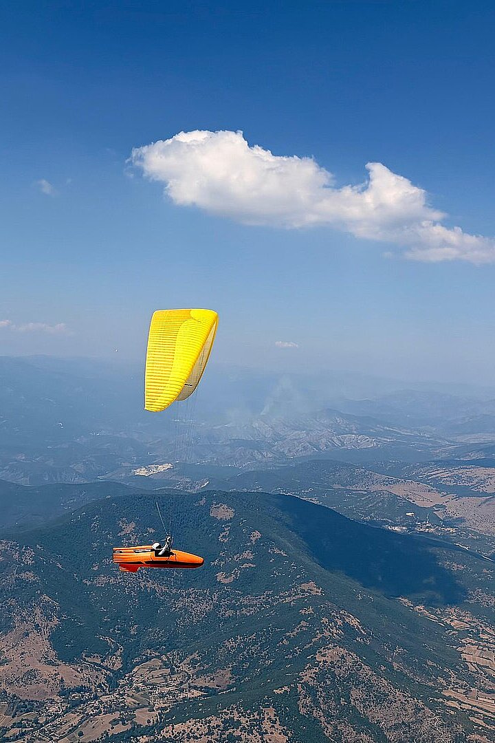 Ein gelber Gleitschirm und ein motorisiertes Fluggerät fliegen über bewaldete Berge.
