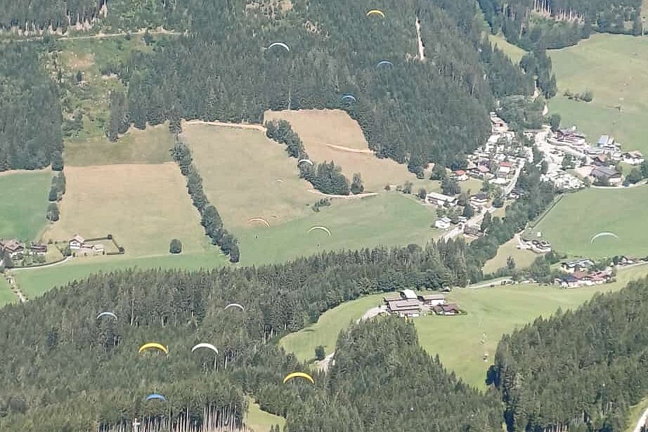 Mehrere Gleitschirme über bewaldeten Hängen und einem Tal mit verstreuten Häusern