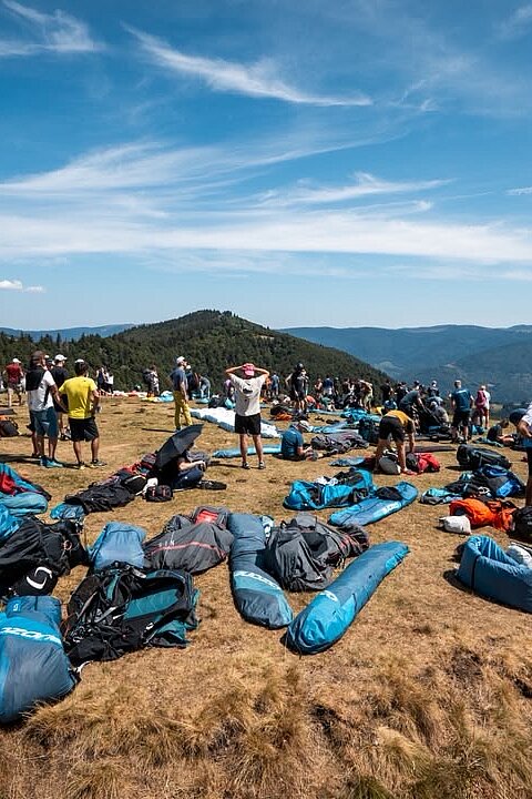 Viele Gleitschirmpakete und Teilnehmer warten auf einer Bergwiese am Startplatz.
