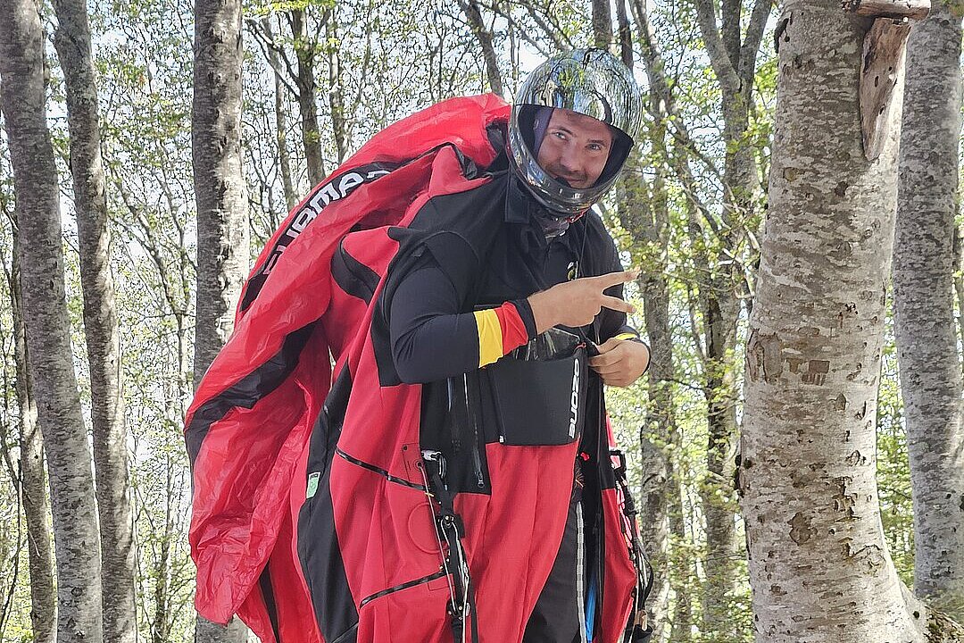 Ein Pilot steht mit Helm und rotem Gleitschirmrucksack im Wald und zeigt ein Peace-Zeichen.