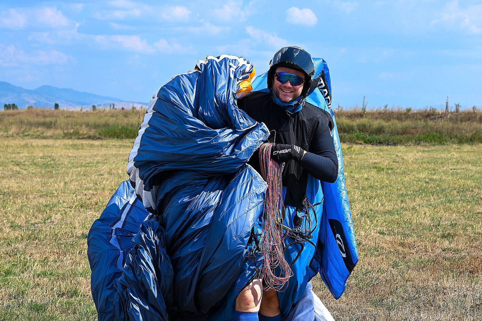 Mann in Helm und Sonnenbrille hält zusammengelegten blauen Gleitschirm auf einer Wiese.