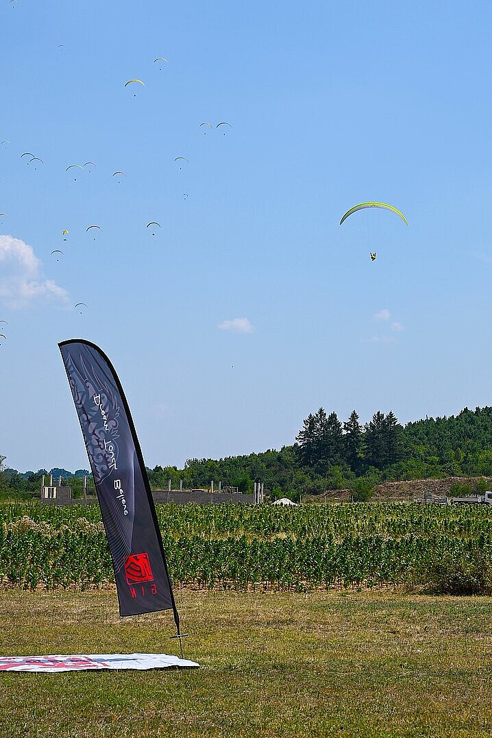 Mehrere Gleitschirme schweben am Himmel über einem Feld mit Banner im Vordergrund.