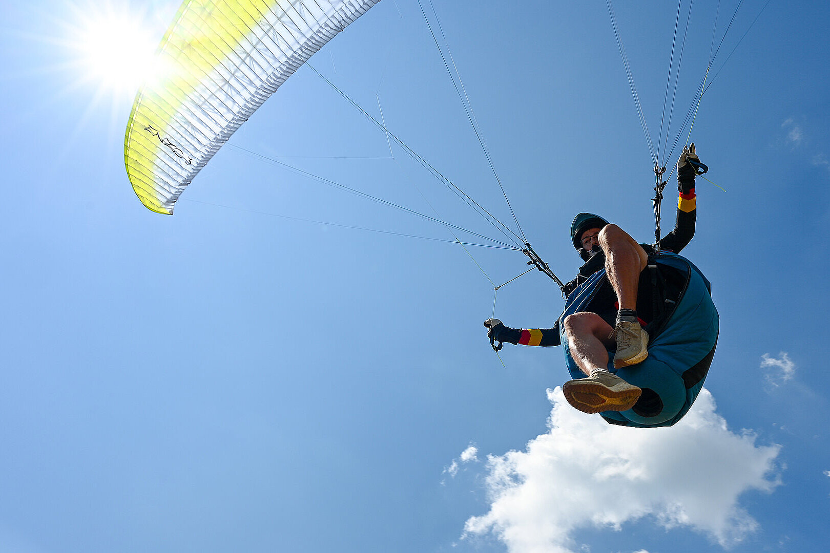Nahaufnahme eines Gleitschirmfliegers beim Start unter blauem Himmel mit Sonne und einzelnen Wolken.