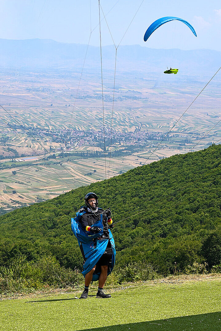 Ein Gleitschirm fliegt über einem Tal mit Feldern und Hügeln.
