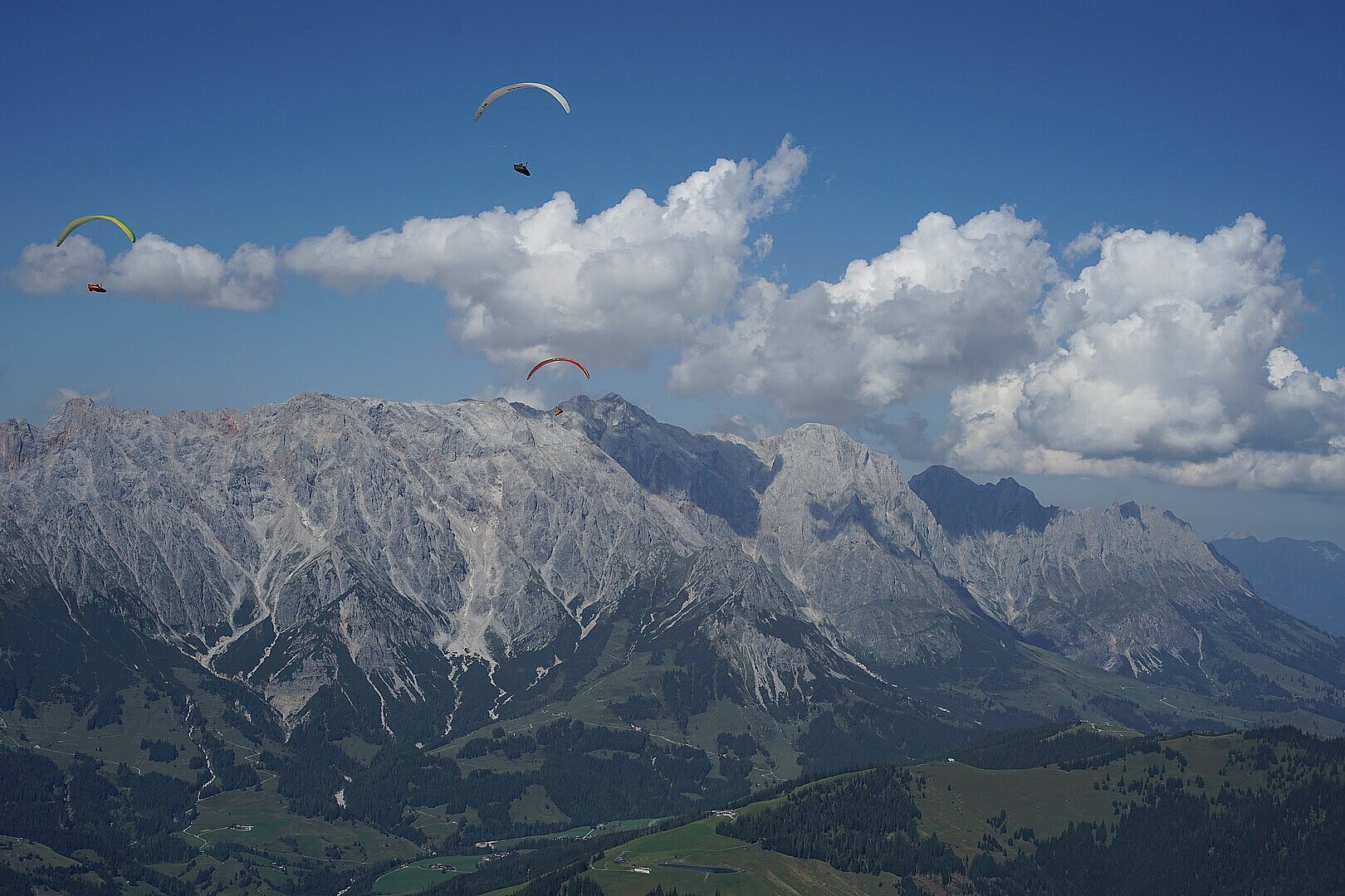 Mehrere Gleitschirmflieger schweben über einer Berglandschaft mit großen Wolken.