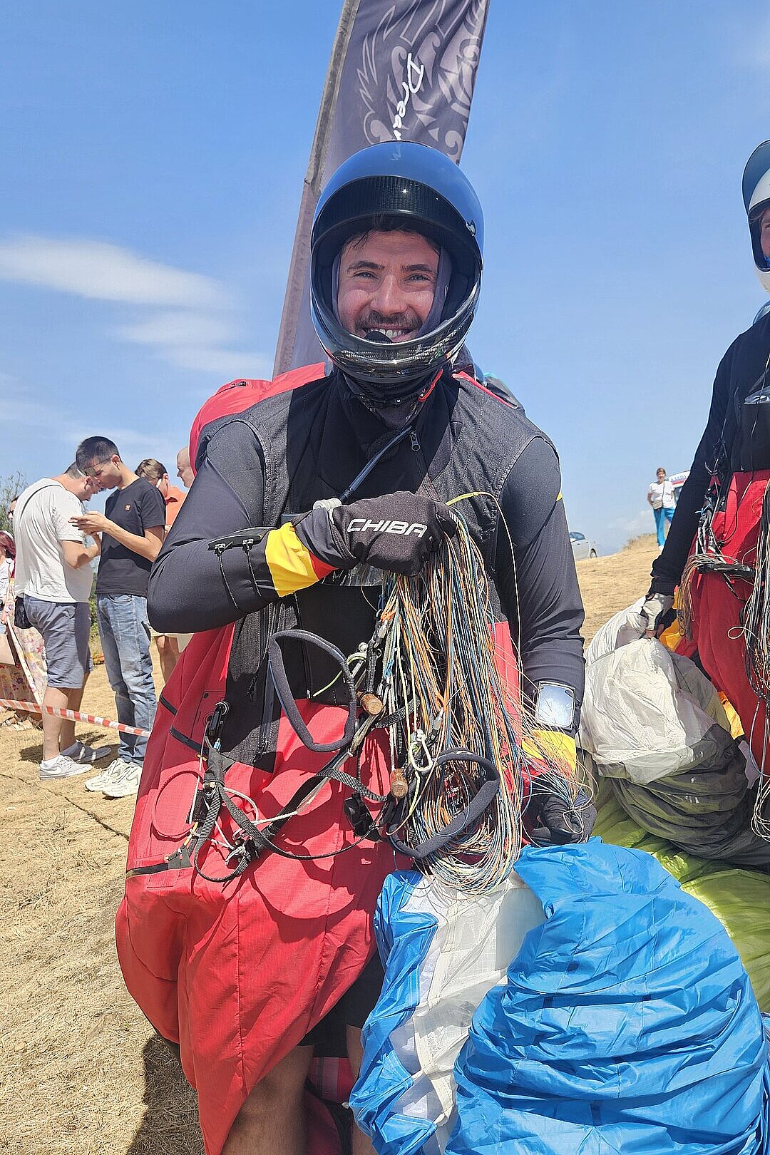 Pilotin oder Pilot mit rotem Gurtzeug und Helm hält Leinen und schaut in die Kamera.