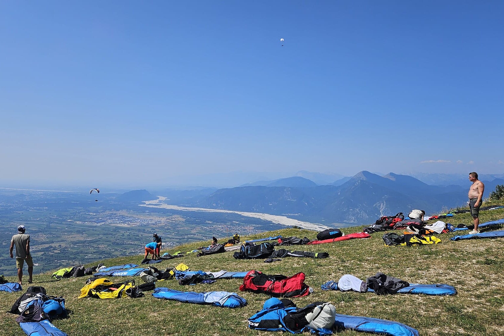 Mehrere Gleitschirme liegen auf einer Wiese am Hang, einzelne Piloten stehen daneben.