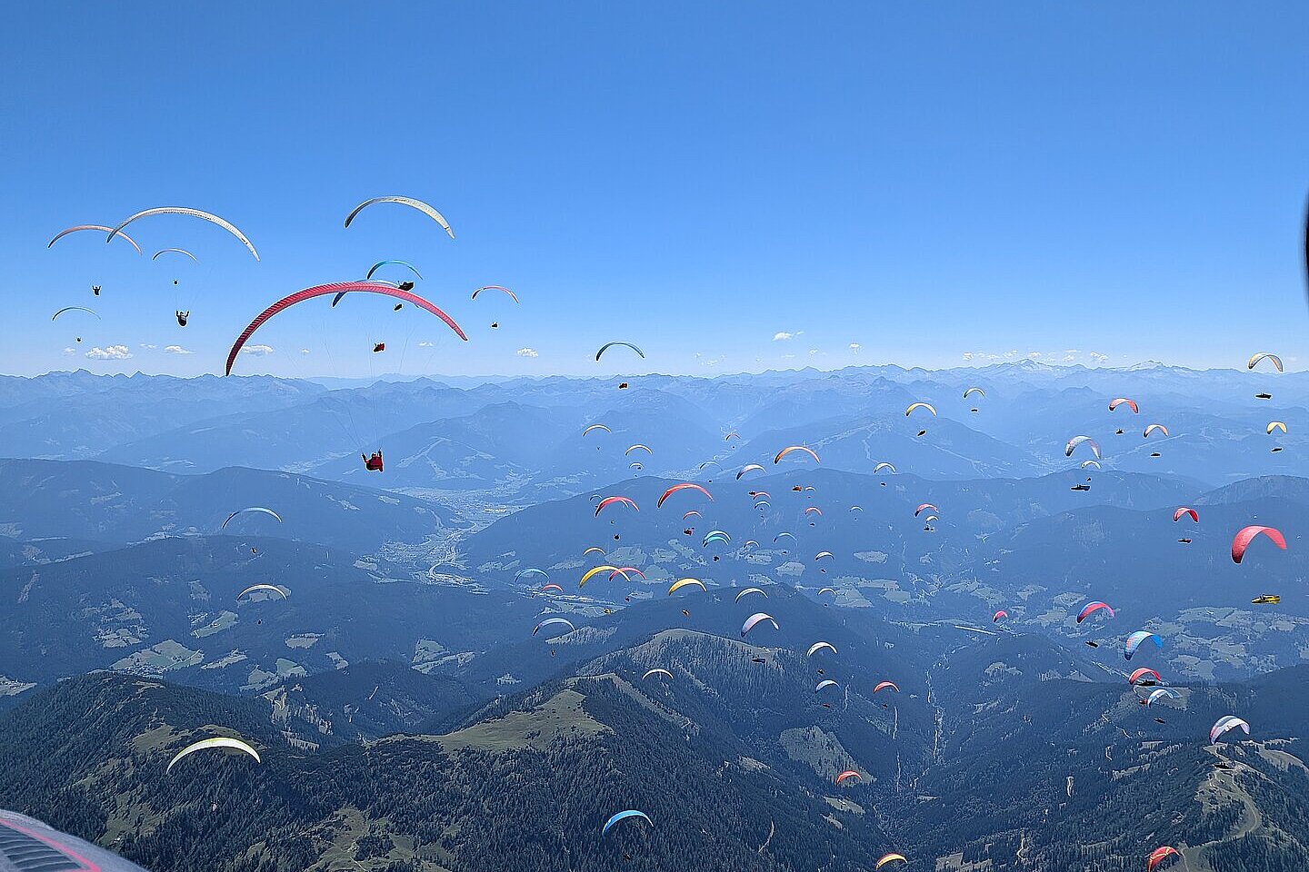 Viele Gleitschirme fliegen über einer Berglandschaft mit Tälern und weit entfernten Hügeln.