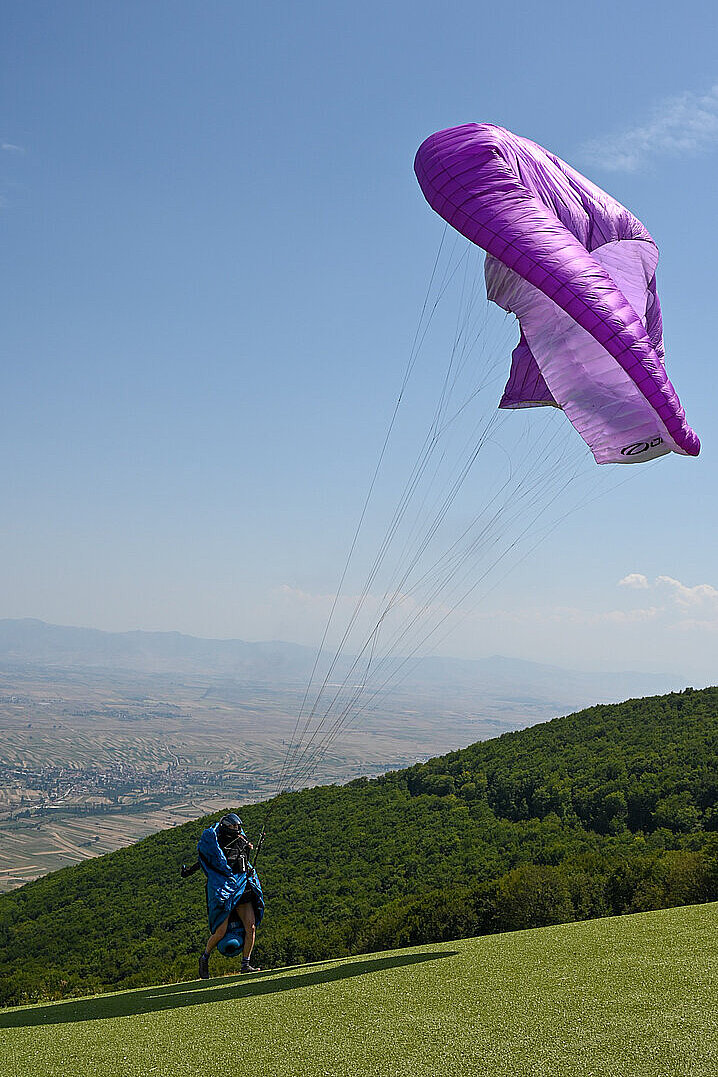 Pilot startet auf einer Wiese mit violettem Gleitschirm vor einem grünen Hügel.