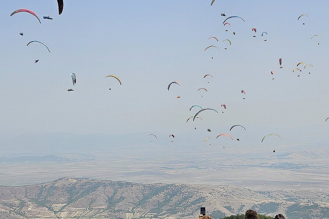 Viele Gleitschirme fliegen über einer weiten Landschaft, drei Personen schauen vom Startplatz zu.