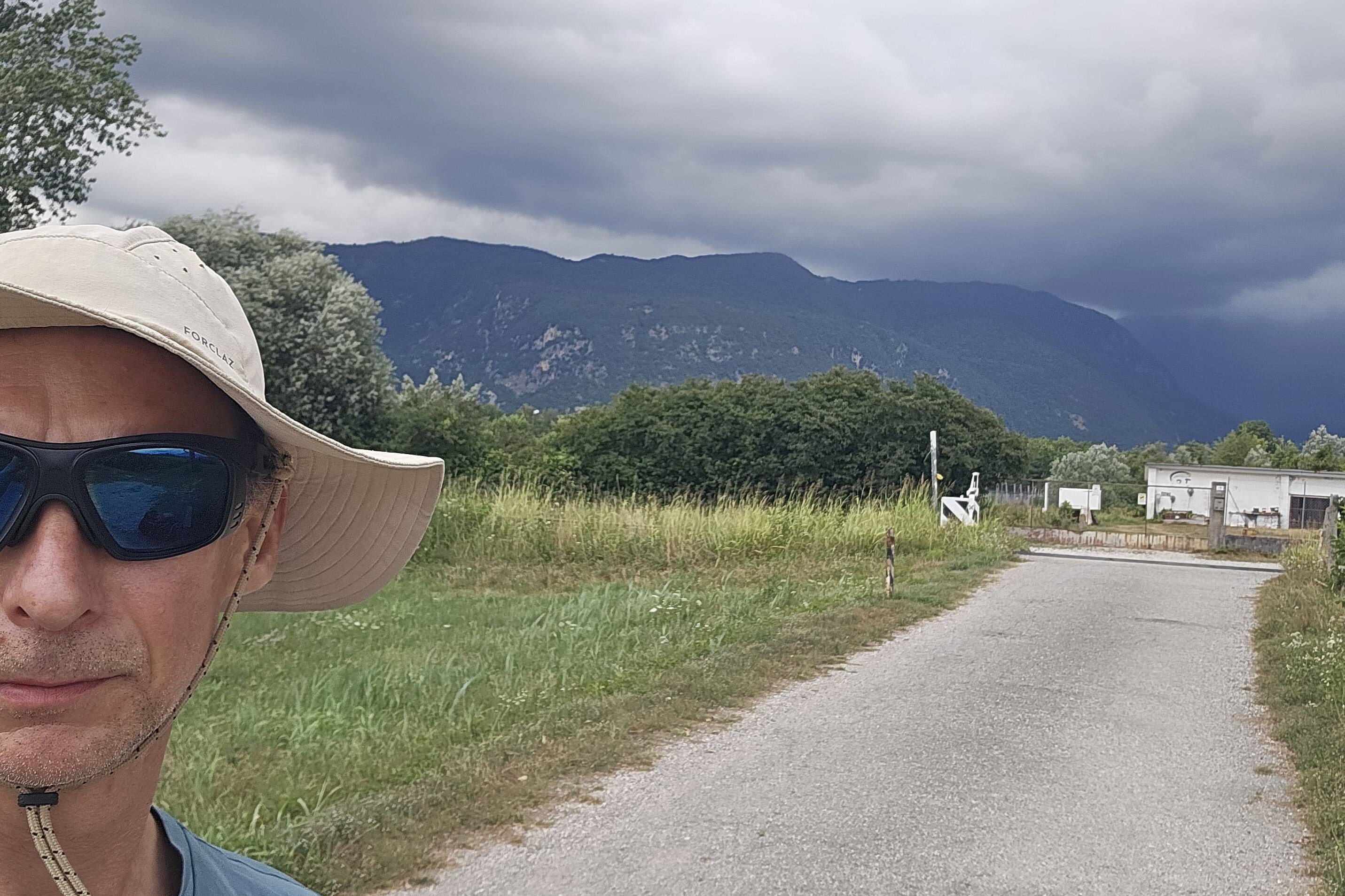 Mann mit Hut und Sonnenbrille macht ein Selfie vor einem Feldweg, Bergen und dunklen Wolken.