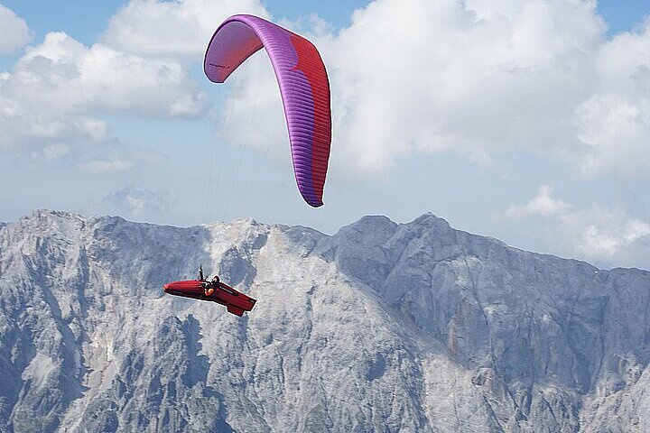 Ein Gleitschirmflieger mit violett-rotem Schirm fliegt vor einer Felskulisse.