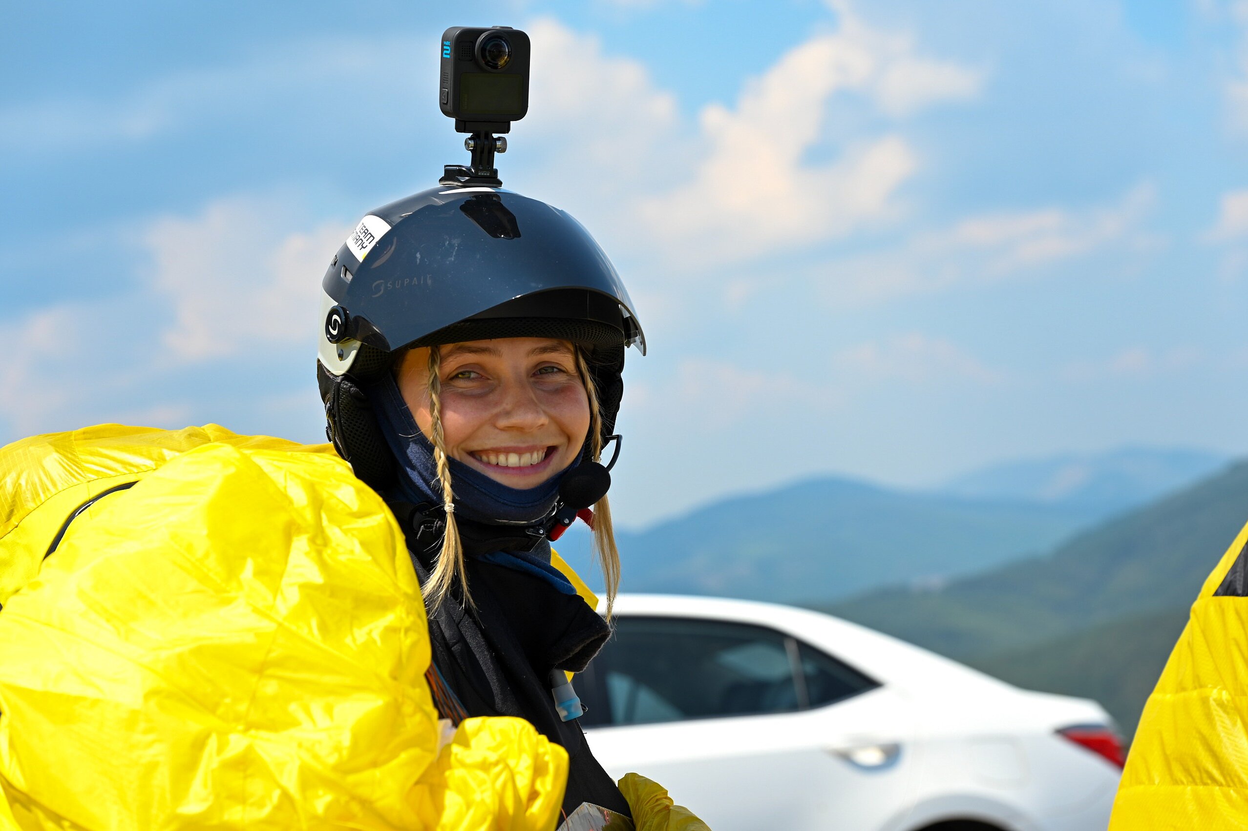 Pilotin mit Helm und Actionkamera lächelt am Startplatz vor Bergkulisse.