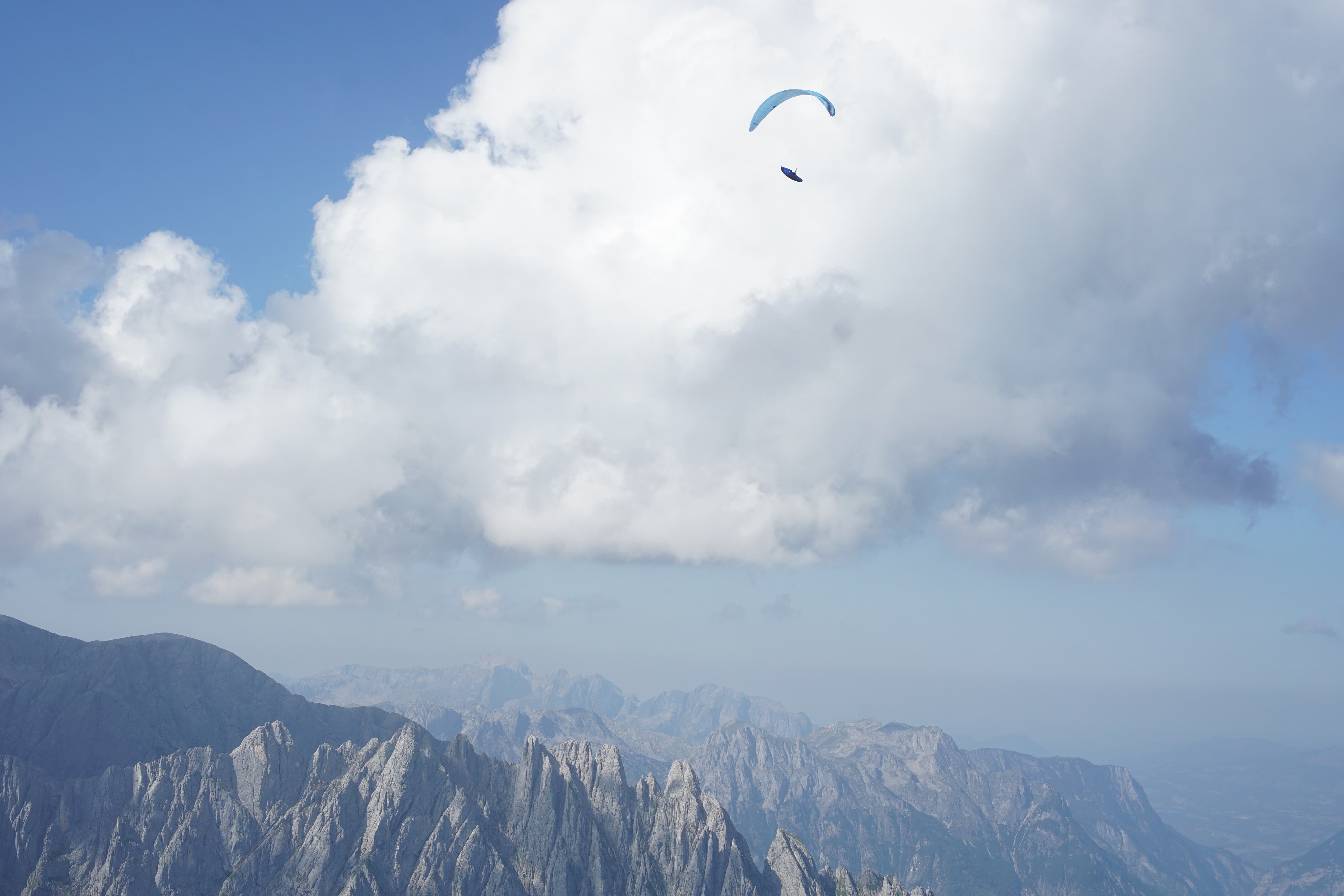 Ein Gleitschirmflieger hängt hoch über schroffen Bergen unter einer großen Wolke.