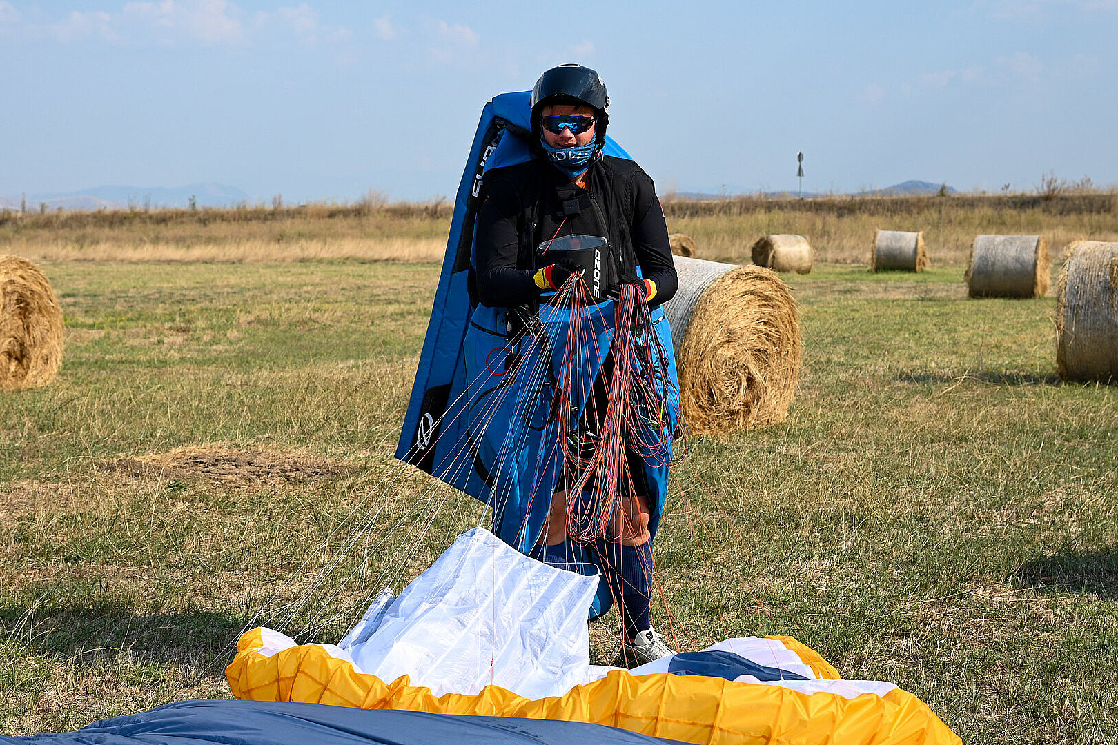 Pilot mit Flugsack und Leinen steht auf einer Wiese neben Heuballen.