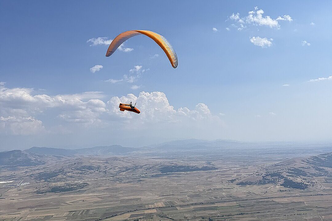 Ein orangefarbener Gleitschirm fliegt über ein weites Tal mit Feldern.