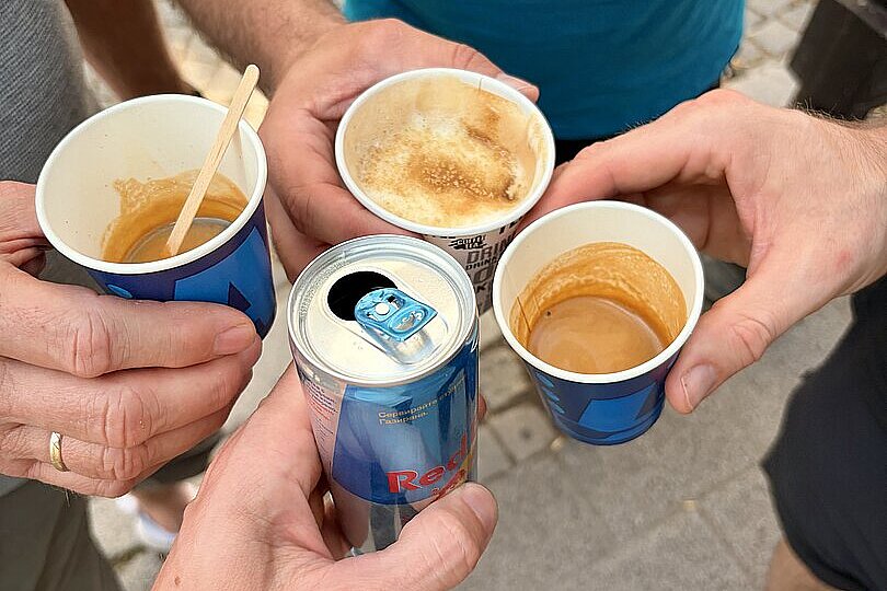 Mehrere Hände halten kleine Kaffeebecher und eine Red-Bull-Dose im Kreis.