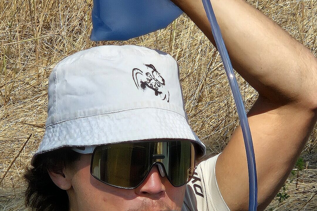 Ein Pilot mit Flasche und Schlauch im Mund sitzt im trockenen Gras und trinkt.