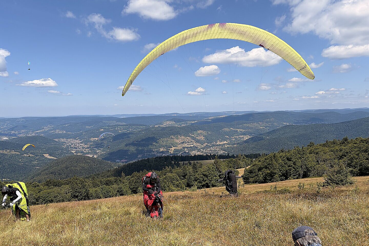Mehrere Gleitschirme starten auf einer Wiese am Hang, im Hintergrund weite Berglandschaft.