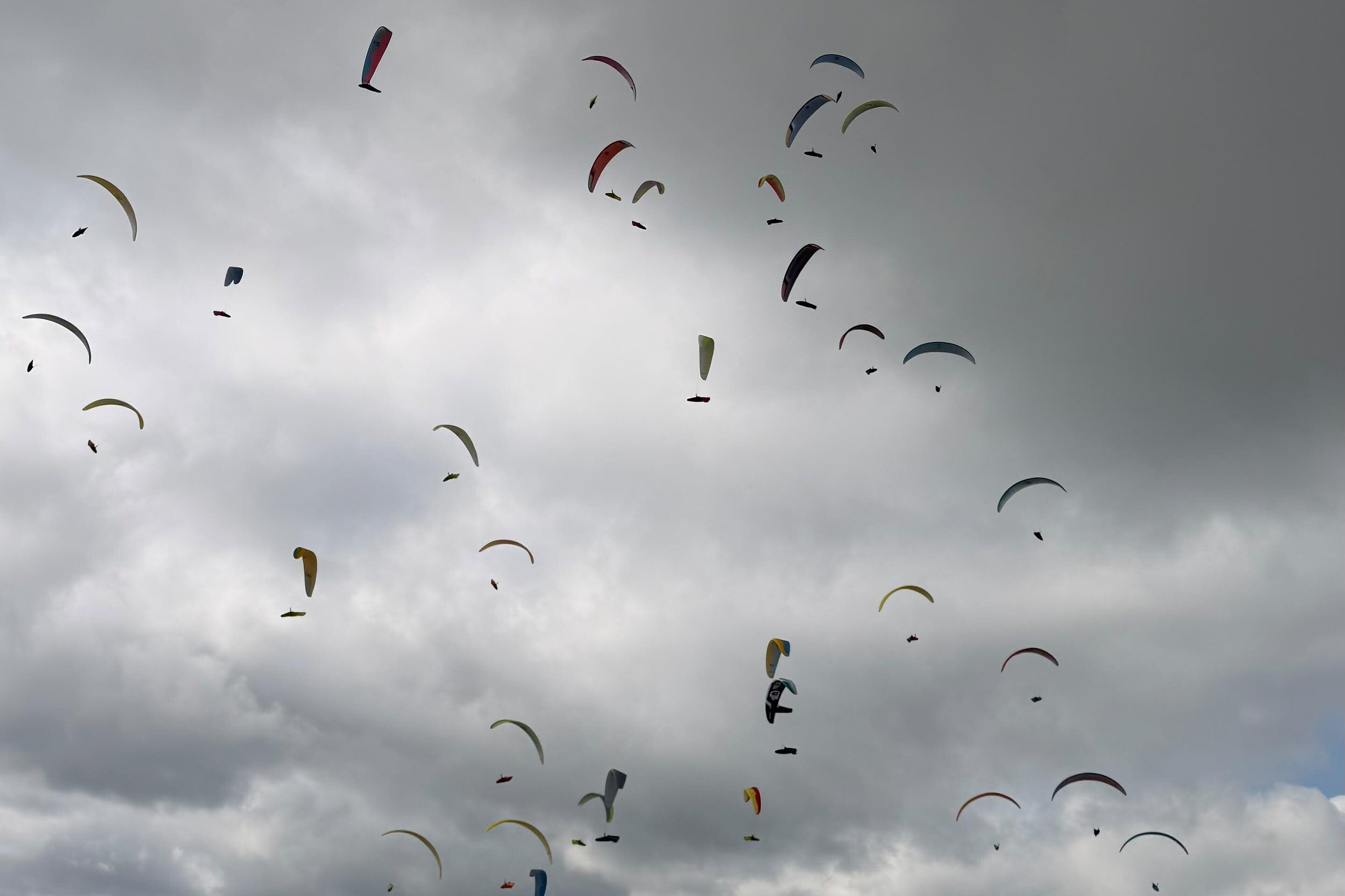 Zahlreiche Gleitschirme schweben über Bergen, einem Tal und Feldern unter dunklen Wolken.