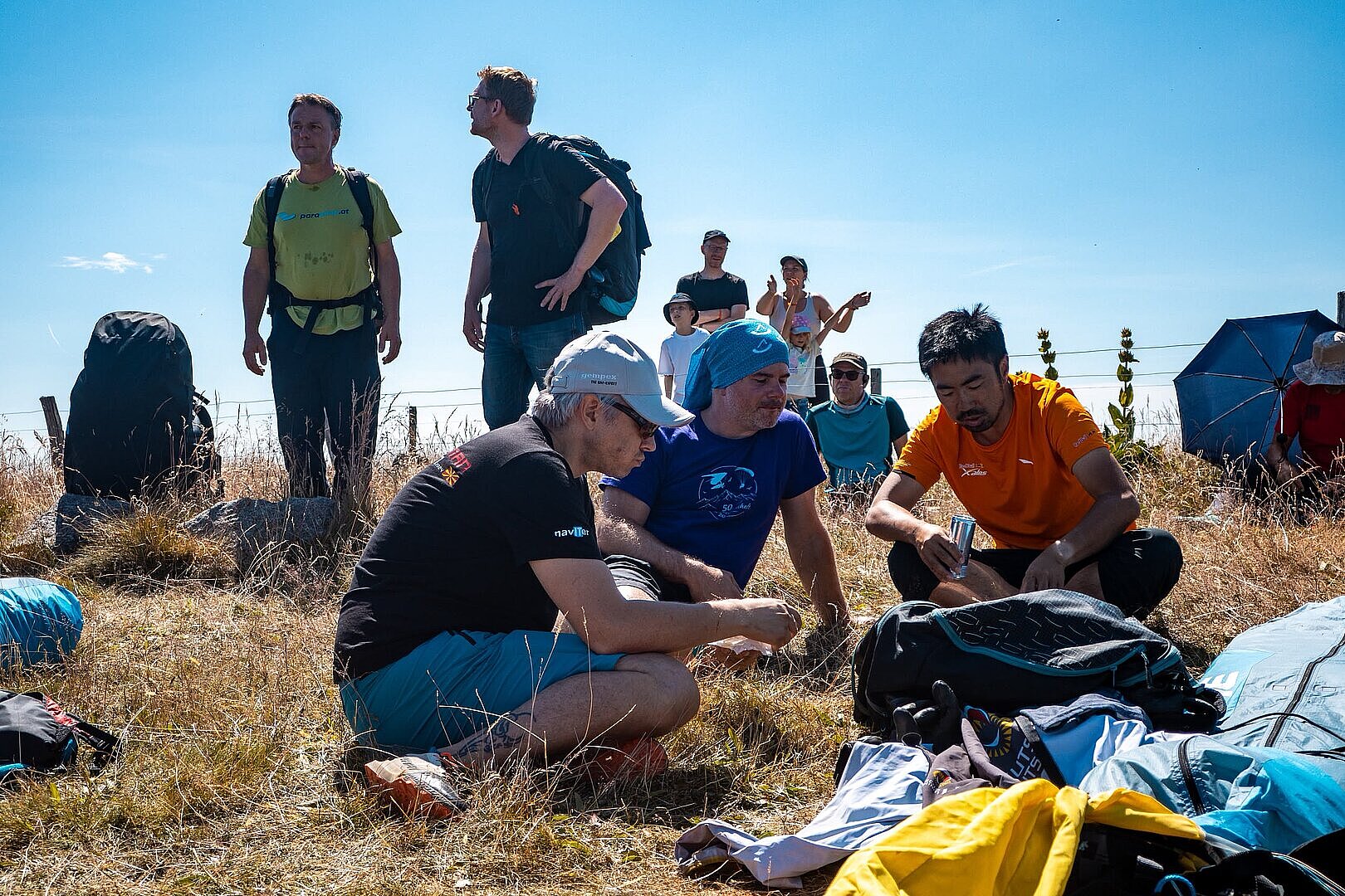 Mehrere Personen sitzen im Gras neben ausgebreiteten Gleitschirmen am Startplatz.