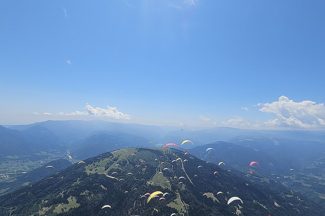 Viele Gleitschirme fliegen über bewaldeten Bergen und einem Tal unter blauem Himmel.