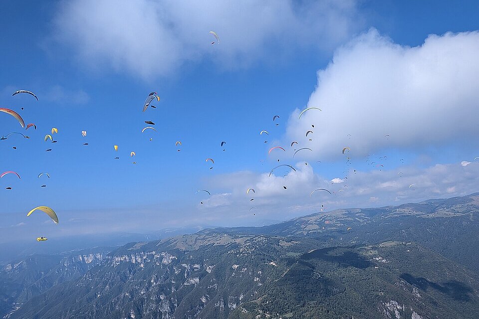 Viele Gleitschirme schweben über einer Berglandschaft mit Tal und Wolken.