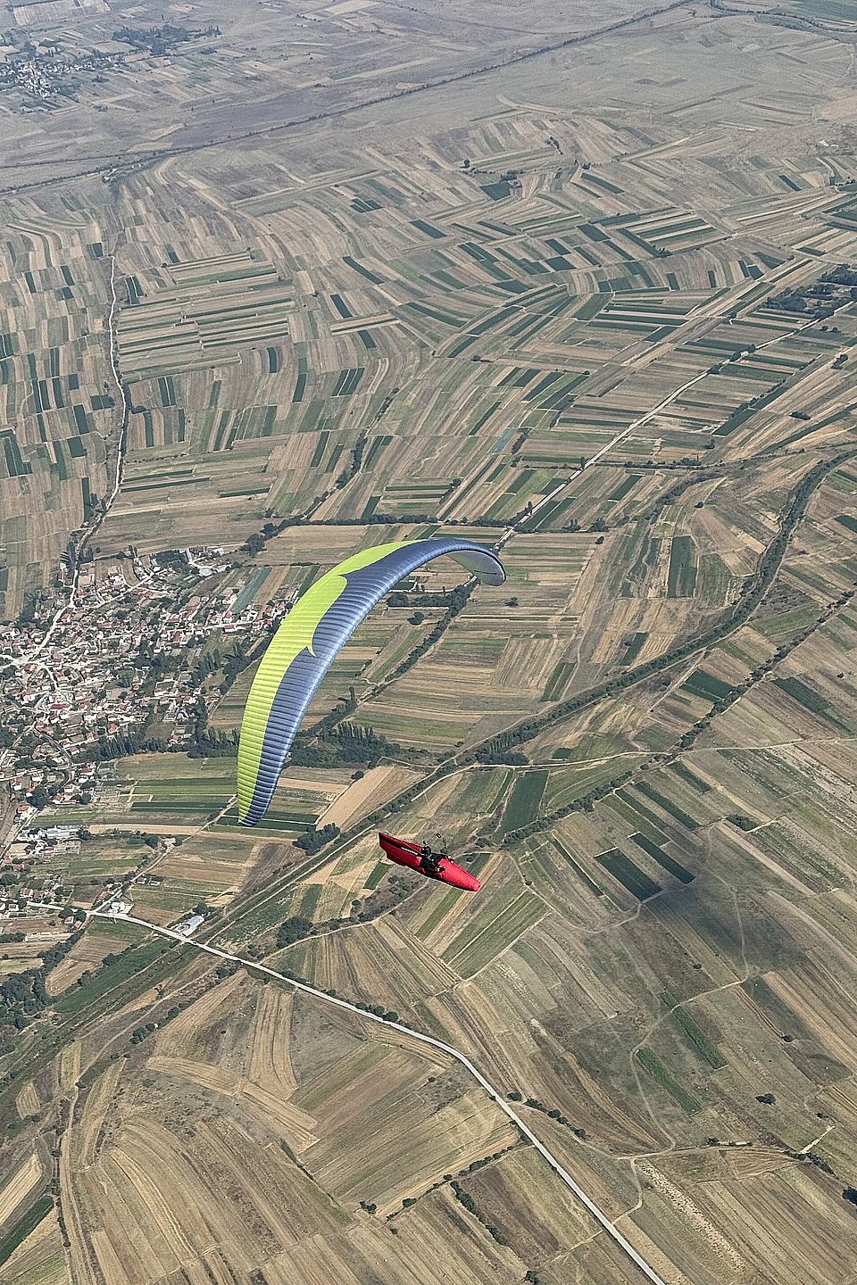 Ein roter Gleitschirm fliegt über ein Mosaik aus Feldern neben einem Ort.