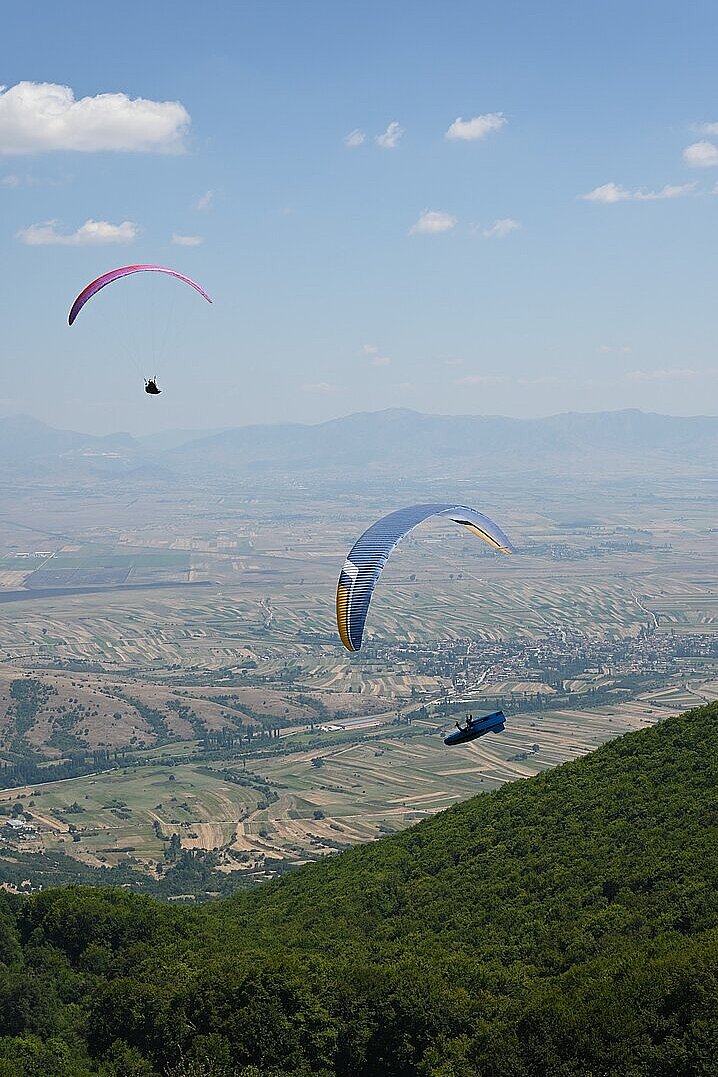 Zwei Gleitschirme fliegen über ein Tal mit Feldern und einem bewaldeten Hügel.