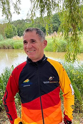 Sportler im schwarz-rot-gelben Teamoutfit vor natürlicher Flusslandschaft