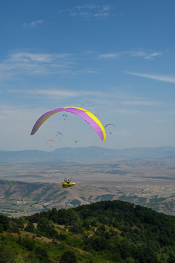 Mehrere Gleitschirme fliegen über einem Tal, einer mit gelb-violettem Schirm im Vordergrund.